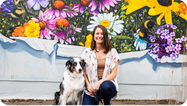 A woman kneels next to her dog, in front of a mural of wild flowers